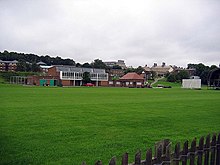 A cricket pavilion joined to a more modern building behind green playing fields, with the sight screens of a cricket pitch to the right