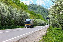 Photographie depuis un côté de la route. La route est de chaque côté longée d'arbres qui fleurissent, avec un camion qui circule. Sur le côté, une borne kilomètrique affiche le nombre 544.