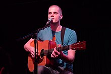 a 20-something-year-old man playing his guitar and singing into the microphone at night.