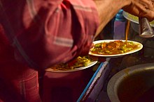 An image of a food vendor preparing masala puri. He has two plates in hand with the dry ingredients of the dish, upon which he pours the spiced gravy.