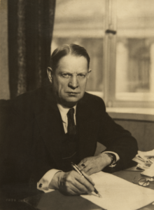 A sepia-toned image of a middle-aged man with a fountain pen sat at a desk.