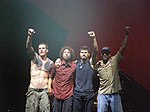 Four men standing, from left to right: a brown-haired shirtless man with tattoos on his shoulders, a man with frizzy brown hair and wearing a reddish shirt, a dark-haired man with a goatee and mustache wearing a sleeveless blue shirt, and a man wearing a visor and a beige shirt. All have draped an arm on another's shoulder, and all but the second to the left have raised an arm in the air.