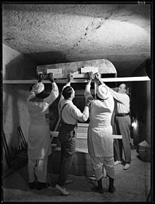 Four men lifting large wooden beams that support a decorated lid