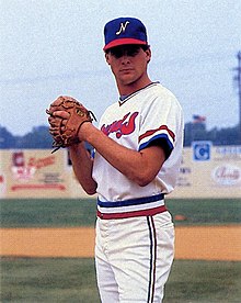 A man in a white baseball uniform and blue cap standing on a field with hands together in his brown leather glove