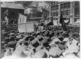 A large crowd of people, some carrying banners. A man in the center is elevated above them and speaking to them