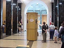 A large hall flanked by black marble columns on the left and right. There is a wooden tower in the middle on which are mounted several plaques. Two people are standing at the tower; one, obscured behind the other, points at a plaque while other people mill about on the sides of the room. The far end opens into a lighted rotunda.