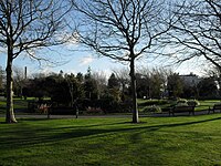 A sunlit park in winter. Behind two bare trees, two benches and lawn in the foreground, a circular path surrounds a slightly sunken area of shrubs with a mound in the centre. Another path leads off diagonally in the left background. In the distance, there is a slim, tall chimney and a large white house with a wide chimneypot.