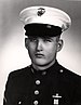 Head and shoulders of a young white man wearing a white peaked cap with black visor and a dark military jacket with bright buttons down the center of the chest.