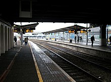 Photo looking along the length of two curved railway platforms, taken from under a shelter and looking out into daylight.