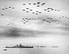 Black and white photo showing a large number of aircraft flying in formation over several World War II-era warships