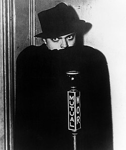 Man in black hat concealing the bottom of his face with a black cape and gazing fiercely. A microphone in front bears the word "Mutual" and the call letters "WOR".