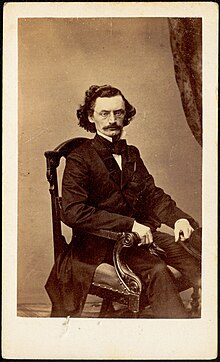 Photograph of Carl Schurz seated in a chair; he has dark hair and a mustache and wears glasses.