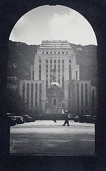 A black and white photo with an arched window framing a view of the front of the HSBC building and the Queen Victoria statue. In the foreground are some parked cars and two pedestrians.