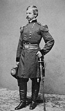 Black and white photo shows a mustachioed man standing. He wears a dark military uniform and holds a sword.
