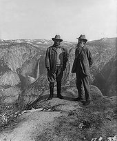Two men stand at a precipice overlooking a valley that has a waterfall in the background.