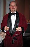 A white man with a goatee is wearing a maroon-colored jacket that ties around the middle with black ribbon. The lapels are wide and black. The jacket is open to the waist, showing a white, high collared shirt beneath with a black bowtie. The man is holding two champagne glasses.