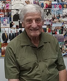 Elderly man with grey hair wearing a khaki-colored shirt sitting at a desk in front of a collage of photographs