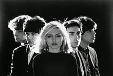 A black and white photo of Blondie posing in a triangular position over a black background.