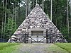 A fieldstone pyramid surrounded by a fence with pine trees in the background