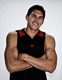 A muscular man wearing a black tanktop with red on the shoulder straps which his arms crossed over his upper abdomen.