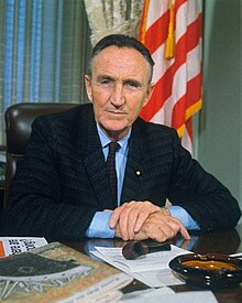 A color photo of Mansfield sitting at his desk in his office, smiling, in 1966.