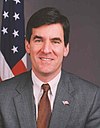 White male with dark hair in a gray suit with an American flag in the background