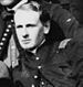 Head of a clean-shaven young man leaning to the side and wearing a military jacket with a single button buttoned near the neck.