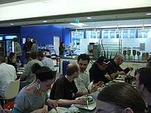 Cette photo en gros plan de l’angle d’une cantine en libre-service montre des gens attablés, une partie de comptoir, des armoires frigos pour les boissons. Le bleu roi des murs domine.