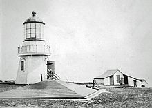 photo noir et blanc avec le phare à gauche des petits bâtiments à droite et la toiture à quatre pans, posée sur le sol, de la citerne à eau