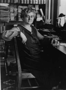 Black and white photograph of a woman in a library, seated at a desk with her arm draped over a chair, holding a pencil in her right hand