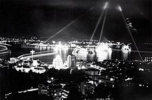 A black and white photo of fireworks at night. The fireworks are shown as streaks of light arching above the water.