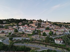 Une photographie du bourg prise de façon à ce que l'ensemble du centre de la commune, pourtant relativement escarpé, soit visible. On y voit une route qui serpente au premier plan, bordée de verdure et surplombée par un grand nombre de maisons individuelles, souvent aux murs beiges et aux toits de briques rouges. Le bâtiment contenant la mairie et l'école du village, sur la gauche, ressort particulièrement. En fond, des bois couvrent la ligne d'horizon.