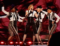 Picture of five women in front of a red background; the one in the center is blonde, has her hair in a braid and sings to a microphone. She's wearing black overalls with a white shirt underneath, and long lace gloves. The rest of the women are dressed in matador outfits.
