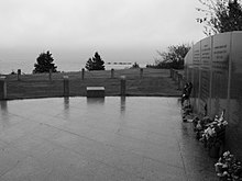 Photo présentant une vue d'un mémorial au bord de la côte. Un sol en pierre précède une vue sur le large et l'océan. À droite, un mur comporte les noms des victimes. Des fleurs sont présentes au pied du mur.