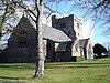 A low cruciform pale-coloured church seen from the southeast with the chancel prominent, beyond which is a broad castellated tower; in the foreground is the trunk of a tree standing in a grassed area