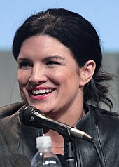 A woman with dark brown hair wearing a leather jacket sits in front of a microphone and smiles at something off-camera.