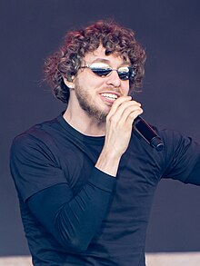 A young man with curly brown hair and facial hair wears reflective sunglasses and two navy blue shirts (a T over a long-sleeved) while holding up a mic.