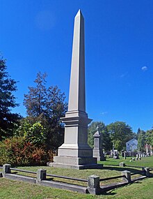 A tall marble obelisk against a clear blue sky with a low stone railing around it. There are smaller monuments, woods and a house in the background.