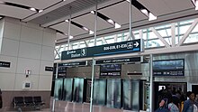 Interior of a modern airport terminal building with glass windows at the top. A sign hanging from the ceiling reads "skytrain Station 3 / D26-D39, E1-E35". Passengers are boarding/alighting a train in the bottom right corner.