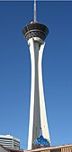 Ground-level view of a tall, concrete tower. The tower curves inward about 30 stories up, but then projects outward. Its uppermost section consists of a rounded, black glass platform and a large, thin spire.