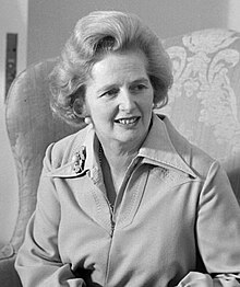 Margaret Thatcher in a jacket with a brooch on a lapel, sitting in an armchair.