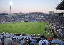 Photo prise depuis une tribune du stade vélodrome de Marseille. Vue sur des supporters de dos au premier plan. Vue sur le terrain et les joueurs au centre de la photo. Vue intégrale de la tribune opposée et vue partielle des tribunes latérales.