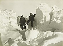Two men in heavy clothing stand surrounded by mounds of ice which extend well above the height of their heads