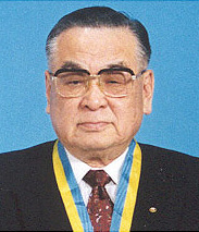 Color photo of an elderly Kawabuchi in a dark suit and tie wearing an award with a blue and yellow ribbon