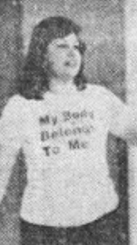 A white woman with dark hair, standing, wearing a t-shirt that says "My Body Belongs to Me"