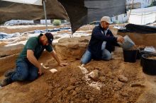 Two people in jeans, shirts and baseball caps knelt down, digging in a trench.