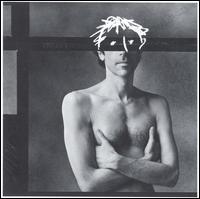 A black-and-white photo of a nude man's bust, wearing an elaborate mask, with his arms folded across his chest