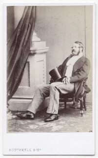 Scarlett with a long mustache and mutton chops sitting in a chair wearing an open black coat and holding a hat. He sits adjacent to a pillar and drape.