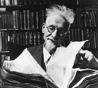 An older white man with glasses, a beard and flyaway hair, looking over a large old book