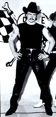 A 23-year-old man stands akimbo, facing forward. He wears a black cowboy hat, sleeveless t-shirt, leather pants, a band on his left wrist and pointed shoes. He has a moustache and has tattoos on his upper arms. He is in front of a sign/art work which is mostly obscured but with a chequered flag, bones and the letters D and E visible. To his left and behind him is another man partly cut off at the edge of the image.
